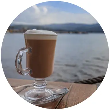 A cappucino on a table with the water, land, and sky behind